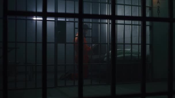 身穿橙色制服的囚犯跪在床边 手持圣经在监牢里向上帝祈祷 狱卒走路 看着罪犯 犯人在狱中服刑 通过金属棒观看 对上帝的信仰 — 图库视频影像