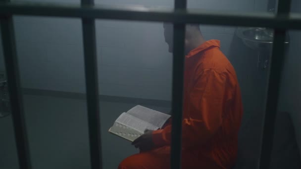 Африканський Американський Язень Помаранчевій Формі Сидить Ліжку Ґратами Читає Біблію — стокове відео