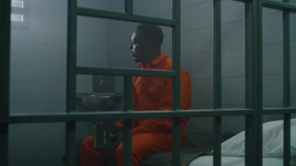 身穿橙色制服的非洲裔美国囚犯坐在狱中的床上 在监狱里阅读圣经 罪犯因犯罪在狱中服刑 拘留中心或教养所 — 图库视频影像