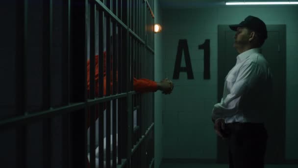 身穿橙色制服的有罪的非洲裔美国人把双手靠在牢房的铁栏上 监狱长在教养所或拘留中心与囚犯交谈 犯人在狱中服刑 — 图库视频影像