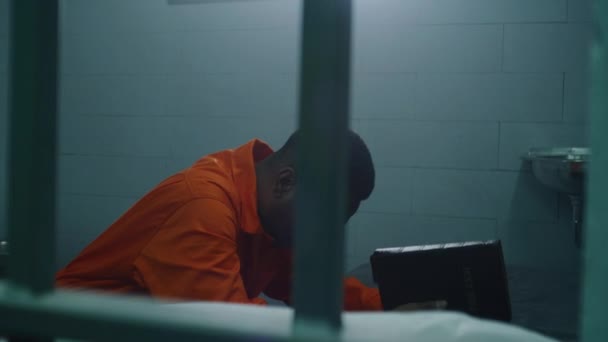 身穿橙色制服的非裔美国宗教囚犯跪在床边 手持圣经向上帝祈祷 男性罪犯看着牢笼的窗户 监狱或拘留中心 对上帝的信仰 — 图库视频影像