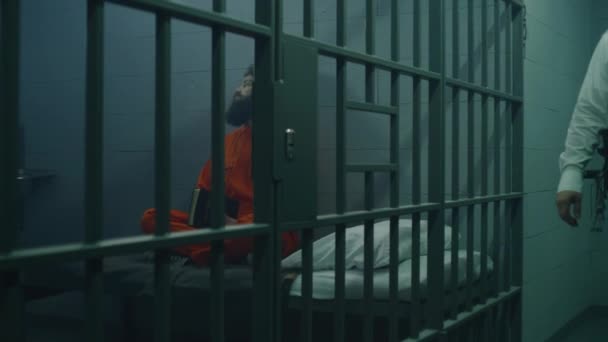 身穿橙色制服的男性囚犯坐在床上 在牢房里阅读圣经 狱警走路 看着罪犯 犯人在狱中服刑 拘留中心或教养所 — 图库视频影像