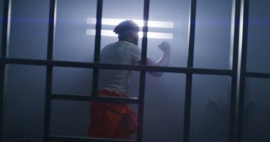 Turuncu üniformalı erkek Afro-Amerikan mahkum hapishane hücresinde gölge boksu yapıyor. Gardiyan hapishane koridorunda yürüyor. Mahkûm, gözaltı merkezinde veya cezaevinde işlediği suçlardan hapis cezasına çarptırıldı.