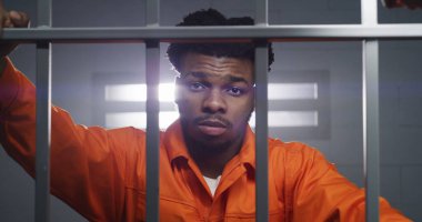 Turuncu üniformalı Afro-Amerikalı bir adam hapishane hücresinde kameraya bakar ve boynu yoğurur. Mahkum hapiste hapis yatar. Islah evinde ya da gözaltı merkezinde bir suçlu. Dikey görünüm.