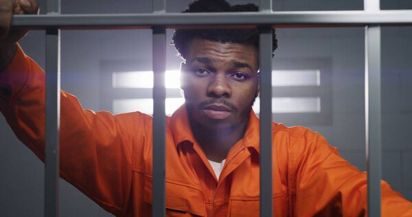 African American man in orange uniform looks at camera and kneads neck in prison cell. Prisoner serves imprisonment term in jail. Criminal in correctional facility or detention center. Portrait view.
