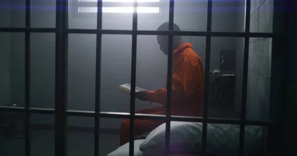 オレンジ色の制服を着たアフリカ系アメリカ人の囚人がベッドに座り 刑務所で聖書を読んでいる 男性犯罪者は 刑務所や拘置所で犯罪の刑期を定めている 神への信仰 バーを通して見る — ストック写真