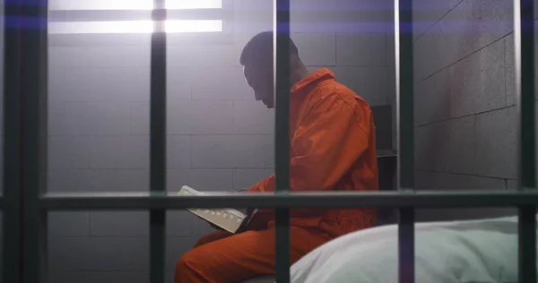 stock image African American prisoner in orange uniform sits on bed behind bars, reads Bible in prison cell. Male criminal serves imprisonment term for crime in jail. Detention center or correctional facility.