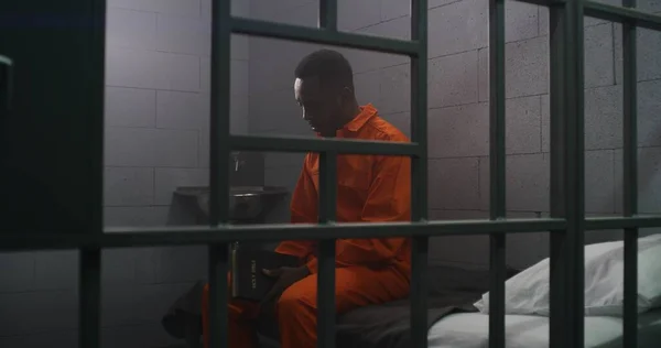 身穿橙色制服的非洲裔美国囚犯坐在狱中的床上 在监狱里阅读圣经 罪犯因犯罪在狱中服刑 拘留中心或教养所 — 图库照片