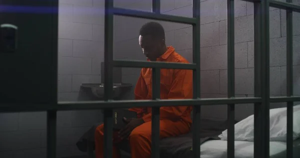 オレンジ色の制服を着たアフリカ系アメリカ人の囚人は バーの後ろのベッドに座り 刑務所で聖書を読んでいます 刑務所では刑務所で有罪判決が下される 拘禁センターまたは矯正施設 — ストック写真