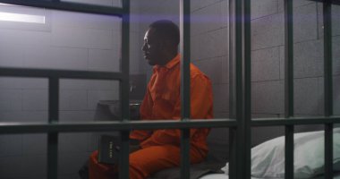Turuncu üniformalı Afro-Amerikan mahkum parmaklıklar arkasında yatıp hücrede İncil okuyor. Suçlu hapiste suçtan hapis cezasına çarptırıldı. Gözaltı merkezi ya da ıslah evi.