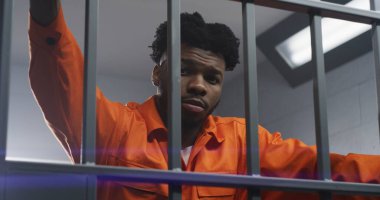 Turuncu üniformalı Afro-Amerikalı adam hapishane parmaklıklarına eğilip kameraya bakıyor. Depresyondaki mahkum hapiste hapis yatıyor. Islah evinde ya da gözaltı merkezinde katil.