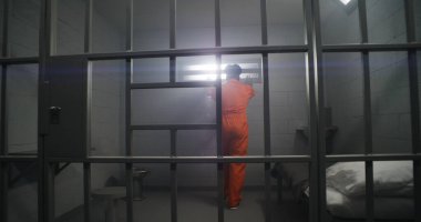Turuncu üniformalı Afrikalı Amerikalı mahkum parmaklıklı pencereye bakar ve hapishane parmaklıklarına yaslanır. Gözaltı merkezinde depresyondaki mahkumlar. Katiller hapis cezasına çarptırıldı.