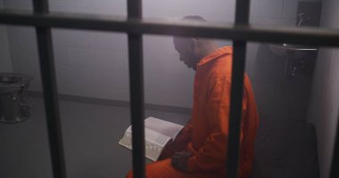 Turuncu üniformalı Afro-Amerikan mahkum parmaklıklar arkasında yatıp hücrede İncil okuyor. Erkek suçlu hapiste suçtan hapis yatıyor. Gözaltı merkezi ya da ıslah evi.