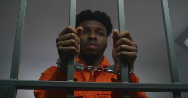 Turuncu üniformalı Afro-Amerikan adam hapishane parmaklıklarında elleri kelepçeli. Depresyondaki katil hapiste hapis yatıyor. Islah evinde ya da gözaltı merkezinde suçlu..