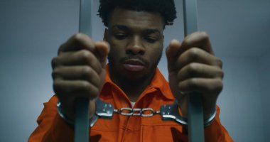Turuncu üniformalı Afrikalı Amerikalı mahkum hapishane parmaklıklarında elleri kelepçeli. Suçlu suçlu hapiste hapis cezasına çarptırıldı. Hüzünlü katil ıslah evinde ya da gözaltı merkezinde.