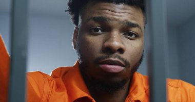 Turuncu üniformalı Afro-Amerikan bir adam hapishane parmaklıklarının arkasında kameraya bakıyor. Mahkum hapiste hapis yatar. Islah evinde ya da gözaltı merkezinde bir suçlu. Portre.