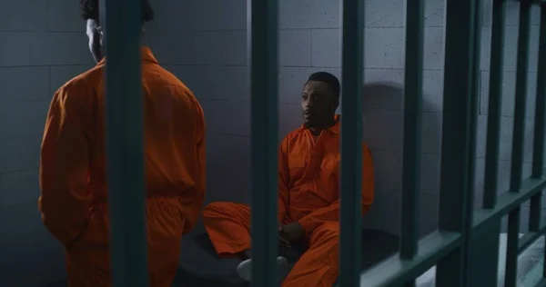 オレンジ色の制服を着た2人のアフリカ系アメリカ人の囚人が話す 一人の男がベッドに座り もう一人は刑務所の中を歩いている 男性は刑務所 拘置所または矯正施設の犯罪のための刑期を務める — ストック写真
