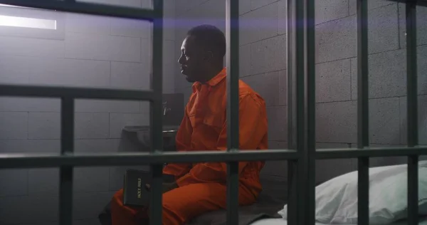 Afroamerykanin Pomarańczowym Mundurze Siedzi Łóżku Kratkami Czyta Biblię Celi Przestępca — Zdjęcie stockowe
