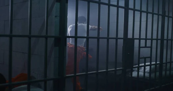 オレンジ色の制服の男性アフリカ系アメリカ人の囚人は 刑務所の監獄でシャドーボクシングをしている ジェイラーは刑務所の廊下を歩いている インメイトは 拘禁施設や矯正施設での犯罪のための刑期を務める — ストック写真