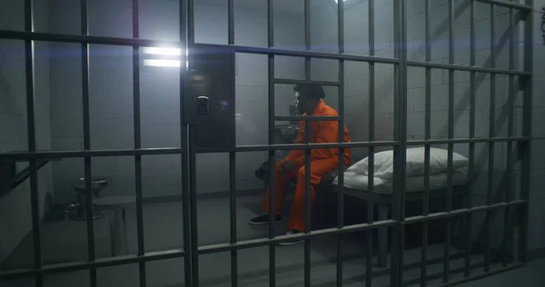 アフリカ系アメリカ人収容所や矯正施設に収容されている オレンジ色の制服を着た抑圧された男は刑務所のベッドに座り 野蛮な窓を見ている 囚人は刑務所の刑務所に投獄される — ストック写真