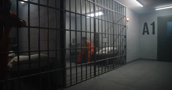 アフリカ系アメリカ人収容所や矯正施設に収容されている オレンジ色の制服を着た抑圧された男は刑務所のベッドに座り 野蛮な窓を見ている 囚人は刑務所の刑務所に投獄される — ストック写真