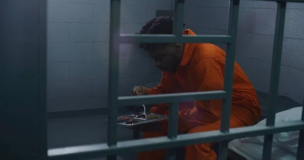 アフリカ系アメリカ人はベッドに座り 夕食を食べ 犯罪のための投獄期間を提供する オレンジ色の制服の囚人は 刑務所の刑務所でトロリーに食事を提供する 刑務所 矯正施設について — ストック写真