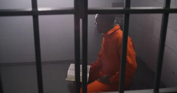 African American Prisoner Orange Uniform Sits Bed Bars Reads Bible — Stock Photo, Image