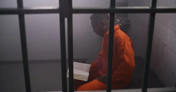 African American Prisoner Orange Uniform Sits Bed Bars Reads Bible — Stock Photo, Image