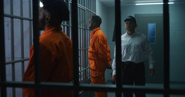 Тюремный работник с полицейской дубинкой гуляет по тюремному коридору и запугивает заключенных. Афроамериканские преступники стоят лицом к камере с руками за спиной. Убийцы отбывают тюремный срок в СИЗО.
