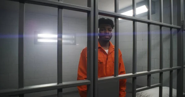 オレンジ色の制服を着たアフリカ系アメリカ人は 刑務所のセルバーに手を貸し カメラを見る 抑圧された囚人は刑務所に投獄される 矯正施設や拘置所での殺人事件 — ストック写真