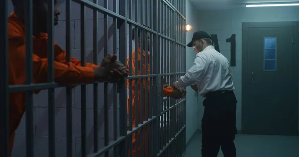 Warden brings new prisoner in jail cell and takes off his handcuffs. African American criminals serve imprisonment term in correctional facility or detention center. Guilty murderers in prison cells.