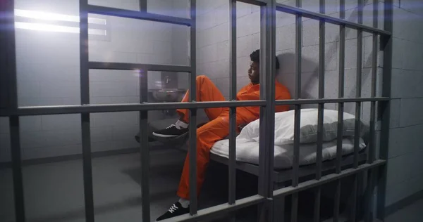 2人の近所の囚人は刑務所の刑務所に収監されている オレンジ色の制服を着たアフリカ系アメリカ人の犯罪者は 刑務所のベッドに座り 野蛮な窓を見ている 拘置所のグロミー収容者 — ストック写真
