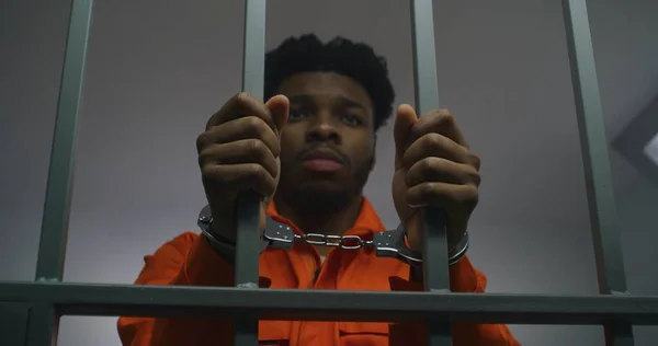 African American man in orange uniform keeps hands in handcuffs on jail cell bars. Depressed murderer serves imprisonment term in prison. Guilty prisoner in correctional facility or detention center.