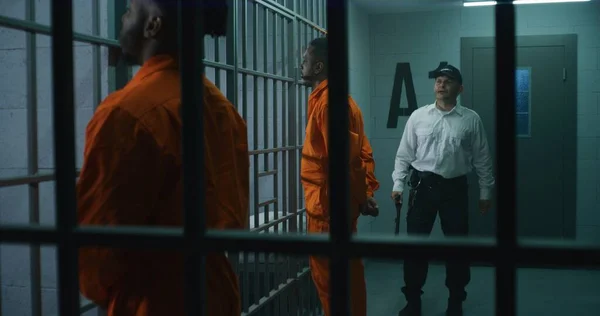 警察のバトンを持つ刑務所の従業員は刑務所の廊下を歩き 囚人をいじめます アフリカ系アメリカ人犯罪者は背後に手で携帯する 拘置所で有罪判決を受けた殺人者 — ストック写真