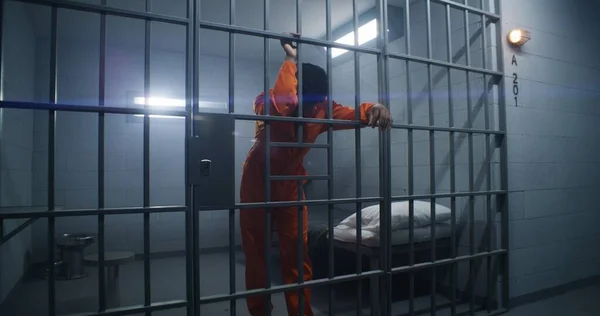 オレンジ色の制服を着たアフリカ系アメリカ人捕虜が刑務所の刑務所のバーに逃げ込み 周りを見回している 有罪のギャングスターは 矯正施設で投獄期間を提供しています 収容所に収容されているグロミー — ストック写真
