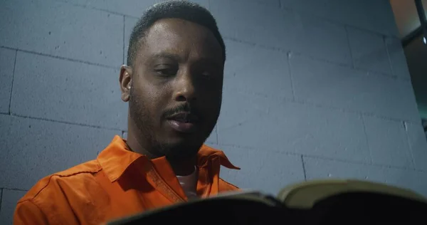 オレンジ色の制服を着たアフリカ系アメリカ人が刑務所に座り 聖書を読んでいます 男性犯罪者は刑務所で有罪判決を受けた 収容所や矯正施設へのオファー ポートレイト — ストック写真
