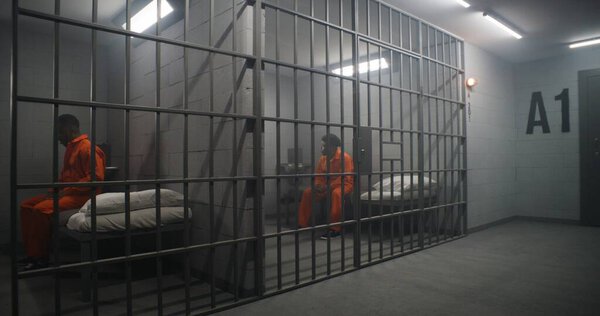 African American inmates in detention center or correctional facility. Depressed man in orange uniform sits on prison bed and looks at barred window. Prisoners serve imprisonment term in jail cell.