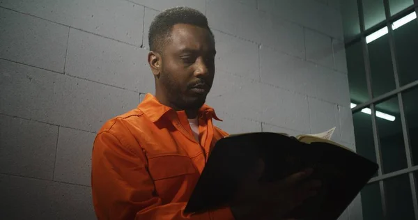 阅读圣经时 身穿橙色制服的非洲裔美国囚犯坐在监牢里 男性罪犯因犯罪在狱中服刑 在拘留中心或教养所的侵犯者 肖像画 — 图库照片