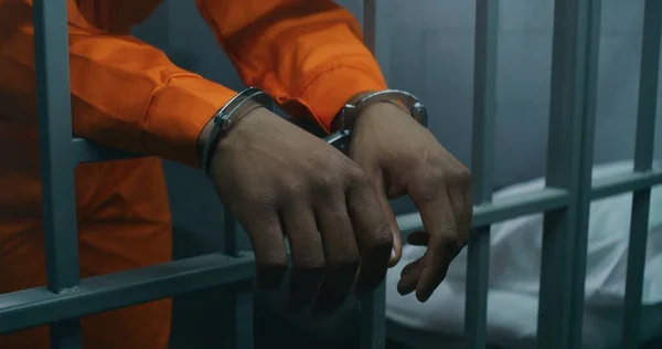 stock image Close up shot of hands in handcuffs leaning on prison cell bars. Criminal serves imprisonment term in correctional facility or detention center. African American prisoner in orange uniform in jail.