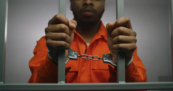 African American prisoner in orange uniform keeps hands in handcuffs on jail cell bars. Guilty criminal serves imprisonment term in prison. Sad murderer in correctional facility or detention center.
