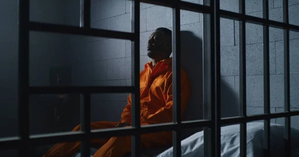 stock image Guilty African American man in orange uniform sits on prison bed and thinks about freedom. Gloomy criminal in correctional facility or detention center. Prisoner serves imprisonment term in jail cell.
