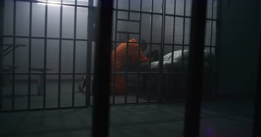 Turuncu üniformalı Afrikalı Amerikalı mahkum yatağın yanında diz çöküp hücrede İncil 'le Tanrı' ya dua ediyor. Erkek suçlu ya hapiste ya da ıslah evinde hapis yatıyor. Metal çubuklardan bak.