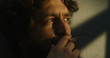 Turuncu üniformalı bunalımlı bir mahkum hapishane hücresinde oturup sigara içiyor. Suçlu, mahkum hapiste suçtan hapis cezasına çarptırıldı. Gözaltı merkezi ya da ıslah evi. Kapatın. Portre.