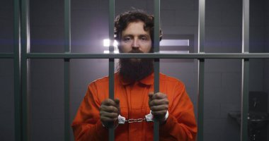 Turuncu üniformalı mahkum metal parmaklıklar tutuyor, hücrede kelepçeli duruyor, kameraya bakıyor. Suçlu hapiste suçtan hapis cezasına çarptırıldı. Gözaltı merkezinde depresyondaki bir mahkum. Portre.