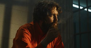 Turuncu üniformalı bunalımlı bir mahkum hapishane hücresinde yatağında sigara içiyor. Suçlu, mahkum hapiste suçtan hapis cezasına çarptırıldı. Güneş parmaklıklı pencereden parlıyor. Cezaevi.