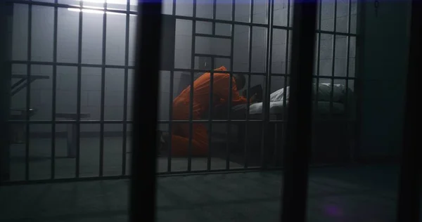 オレンジ色の制服のアフリカ系アメリカ人の囚人がベッドの近くでひざまずき 聖書の刑務所で神に祈ります 男性犯罪者は刑務所や矯正施設で投獄期間を過ごしている 金属棒を通して見る — ストック写真