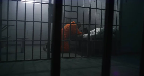 身穿橙色制服的非裔美国囚犯跪在床边 用圣经在监牢里向上帝祈祷 男性罪犯在监狱或教养所服刑 透过金属棒观看 — 图库照片