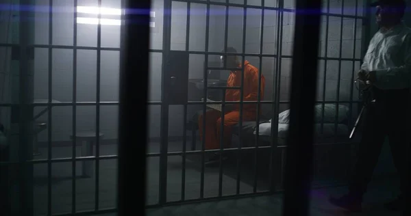 オレンジ色の制服を着たアフリカ系アメリカ人の囚人がベッドに座り 刑務所で聖書を読んでいる 刑務所の警官が歩いて 犯罪者を見張る インメイトは刑務所や拘置所で犯罪の刑期を務める — ストック写真