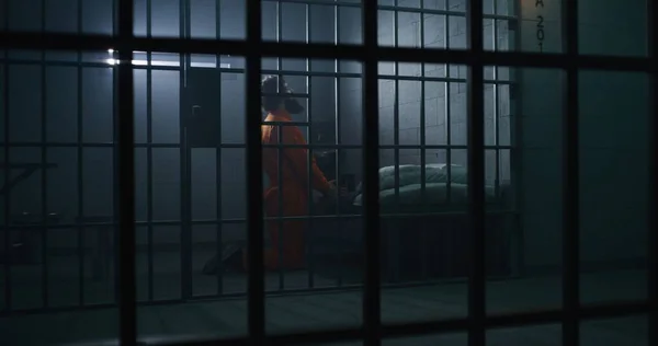 ベッドの近くでオレンジ色の制服を着た囚人は 聖書を保持する刑務所で神に祈ります ジャイラーが歩いて 犯罪者を見張る インメイトは刑務所で投獄期間を過ごす 金属棒を通して見る 神への信仰 — ストック写真