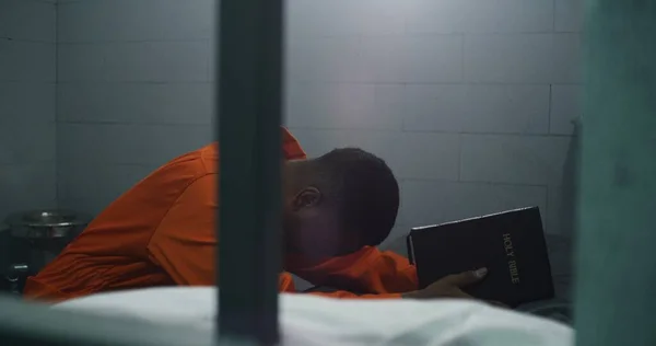身穿橙色制服的非洲裔美国宗教囚犯跪在床边 手持圣经在监牢里向上帝祈祷 男性罪犯在监狱或拘留中心服刑 对上帝的信仰 — 图库照片
